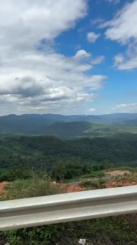 The video begins by looking over a guardrail at a lush green mountainous landscape under a bright blue sky with fluffy white clouds. The camera then pans right, revealing more of the rolling hills covered in dense forest. In the foreground, there are dry grasses and some red dirt. As the video continues, a small, simple building with a reddish-brown roof appears on the left side, partially obscured by tall grass. The camera keeps moving, showcasing more of the expansive view of the mountains and the sky. A solitary tree with a full canopy stands out against the backdrop of distant hills. Finally, the view shifts to include more trees in the foreground and on the slopes, with the camera continuing its slow pan across the scenic, hilly terrain. The environment appears natural and somewhat undeveloped.