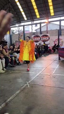 The video captures a vibrant dance performance in an indoor gymnasium-like space.  A woman dressed in a bright yellow dress with orange fringed shawl and red heeled shoes is dancing. A man joins her, and they spin while holding hands. A seated audience surrounds the performers and a person who is part of the audience is filming the video, and their hand is visible in the foreground, clapping along with the others in the audience. The space features a high, arched ceiling with translucent panels for lighting, and walls adorned with posters and logos. The overall atmosphere is celebratory and energetic.
