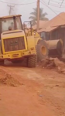 The video shows a yellow CAT bulldozer working on a dirt road in what appears to be a village. The bulldozer is moving forward, pushing dirt and debris with its blade. A person is operating the bulldozer. There are buildings and power lines visible in the background, and a few people can be seen walking in the distance. The scene is dusty and the overall lighting is hazy.