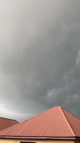 The video captures a slightly overcast sky with a hint of storm clouds gathering. The scene focuses on the rooftops of two houses, one with a reddish-brown corrugated metal roof and the other with a darker, possibly asphalt shingle roof. A tall utility pole with electrical wires stretches diagonally across the frame, its insulators visible. In the foreground, glimpses of green foliage from trees can be seen, suggesting an outdoor setting in a populated area, likely a residential neighborhood in Barakin Sabawa, Nigeria.

The overall mood is subdued, with the graying sky hinting at approaching rain or a change in weather. There are no people visible in the video, and the focus is on the architecture and the atmospheric conditions. The time of day appears to be late afternoon or early evening, given the diffused lighting and the darkening clouds. The scene is static, with no discernible activity other than the slow pans and zooms that transition between different parts of the sky and the houses. The prominent elements are the stark lines of the rooftops against the vast expanse of the sky and the industrial presence of the utility pole.