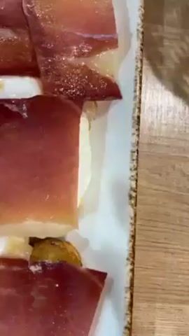 The video shows a table with two plates of food and a glass of red wine.  One plate contains slices of cured ham on top of what appears to be fried potatoes and eggs. The other plate holds fried squid and what seems to be fried fish, garnished with lemon wedges.  A hand is seen pouring red wine into a glass.  The table surface is wood-toned, and a cell phone is also visible on the table. There seems to be a lively, noisy restaurant atmosphere.