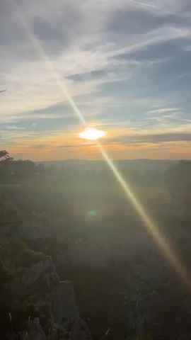 The video opens with a wide shot of a sunlit landscape, possibly during sunrise or sunset, as indicated by the low angle of the sun and the warm hues in the sky. The sun's rays are a prominent feature, casting a diagonal light across the frame and creating a lens flare effect. In the foreground, a rocky cliff face occupies the lower left portion of the screen, with a sparse covering of moss and small plants. The terrain appears rugged and uneven.

As the video progresses, the camera pans slowly across the scene. The midground reveals a hazy valley or plain dotted with trees and some structures that are indistinct due to the atmospheric conditions and distance. A distinct line of trees can be seen on the horizon, separating the sky from the land.

The camera continues to move, revealing more of the cliff's edge and the surrounding vegetation. A path, possibly a hiking trail, becomes visible along the edge of the cliff in the latter part of the video. The vegetation on the cliff top consists mainly of trees, with some grasses and scrub. The overall environment appears to be a natural landscape, likely a mountainous or hilly region. The sky is partly cloudy, with streaks of white clouds against a blue and orange gradient. There are no visible people or animals in the video. The lighting suggests it is either early morning or late evening.