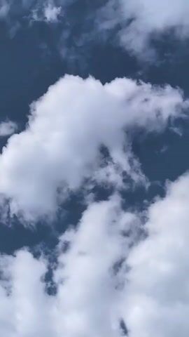 This is a close-up, vertical shot of the sky filled with white, puffy clouds against a deep blue background. The clouds are textured with variations in light and shadow, suggesting depth and movement. Some clouds appear wispy and thin, while others are dense and voluminous. The image is static, with no discernible action or objects other than the clouds and sky. The lighting indicates it is daytime, likely bright sunlight illuminating the clouds.