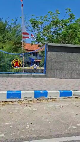 The video shows an exterior view of a university campus. A large sign with the words "UNIVERSITAS MADURA" is visible, along with the university's emblem. There is a banner welcoming new students, featuring pictures of individuals in formal attire. Other elements in the scene include trees, a clear blue sky, a cell tower, and a roadside stall. The overall atmosphere appears to be bright and sunny, suggesting daytime. The footage is captured from a moving vehicle, indicated by the slight blur and panning motion.