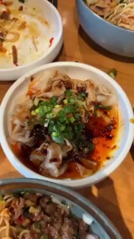 Several plates of food are arranged on a light brown wooden table. One plate is partially eaten, with a fork resting on it. Another plate contains what appears to be dumplings topped with chili oil and chopped green onions. A third, larger bowl contains noodles with some kind of meat and vegetable topping.