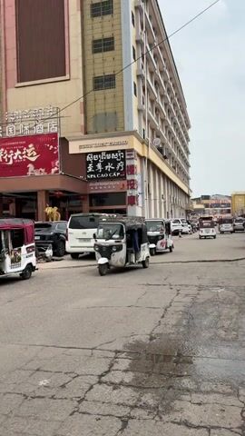 The video shows a street scene in front of a building labeled "Good-Luck Casino". Several white auto-rickshaws, commonly known as "tuk-tuks," are parked or moving slowly on the cracked asphalt street. A white van is parked near the casino entrance. The building is tall and multi-story, with a mix of architectural styles. The sky is overcast, creating a subdued lighting environment. Signs in Chinese and Khmer adorn the building's facade, indicating a possible international or mixed-cultural setting. No people are clearly visible within the tuk-tuks.