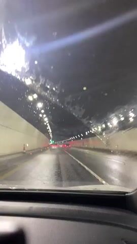 The video shows a point-of-view shot from inside a car driving through a tunnel. The windshield has water droplets and streaks on it, obscuring some of the view. The tunnel is well-lit with rows of lights along the ceiling. The road appears wet, reflecting the lights. Several cars are visible ahead, their headlights and taillights illuminated. The tunnel walls are light-colored and curved, forming an arched ceiling. There are also orange traffic cones visible along the side of the road. The interior of the car is partially visible in the foreground, including the dashboard. The video is brief, lasting only a few seconds, and the sound of traffic is faintly audible.