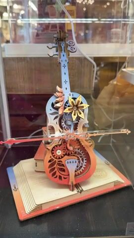 This is a close-up, slightly angled shot of a wooden, mechanical model of a cello. The cello is intricately designed with visible gears and ornate cutouts. It is positioned on top of a wooden book or stand that is open, with stylized pages. The cello itself is decorated with floral elements and a flower shape on the body. A miniature bow is positioned as if to play the cello. The background appears to be a retail display, with glass or acrylic casing and other items visible out of focus. The lighting is bright and highlights the details of the model. The video appears to be a loop, with the cello model remaining stationary throughout.