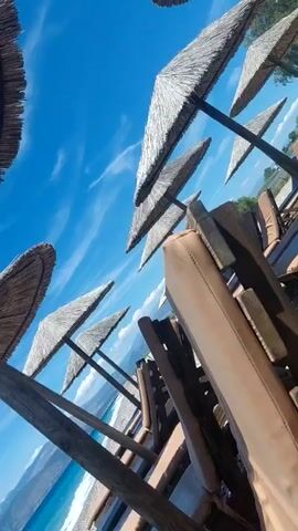 The video shows a view of a beach with several straw umbrellas and lounge chairs in the foreground. The sea is a vibrant blue, with gentle waves breaking on the pebble beach. Mountains are visible in the background under a clear blue sky with some wispy clouds. The camera pans slightly to the left, revealing more of the beach and the coastline. The overall impression is a peaceful and sunny day at the seaside.