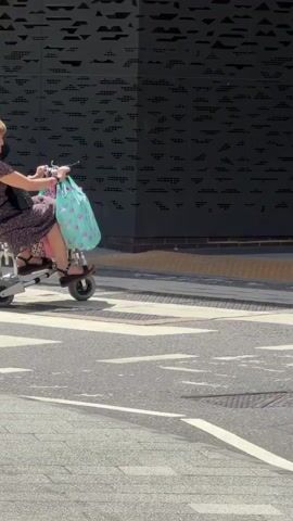 A woman with blonde hair, wearing a dark floral dress, rides a yellow and gray mobility scooter with three wheels. She is carrying a light blue shopping bag with a pink pattern on the handlebars. The scooter is on a paved surface, possibly a sidewalk or a crosswalk, with the woman moving from left to right across the frame. In the background, there is a building with a dark patterned facade and a brown section. A brick wall and a metal railing are also visible. The scene appears to be outdoors during the daytime, with sunlight casting shadows.