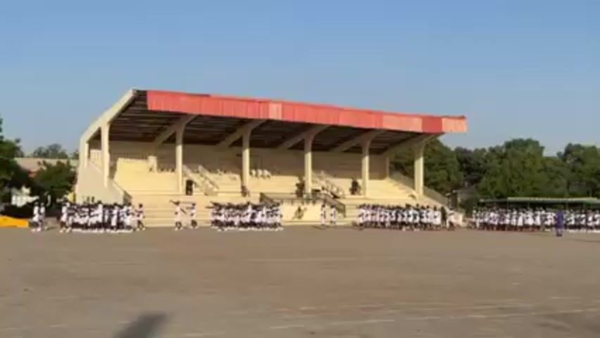 A wide shot shows a crowd of people in uniforms standing on a large dirt field, facing a small stadium with a red roof. The people appear to be in formation and are moving their arms. The sky is blue and the lighting is bright, suggesting a sunny day. Trees line the periphery of the field. The atmosphere seems celebratory or ceremonial. A bird flies across the field in front of the camera.
