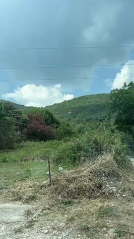 A car drives along a road passing by green trees, hills, and cloudy skies. A white fence is visible in the distance, with a street lamp to the right. The car passes by a concrete wall with blue and green graffiti. A white building is visible on a hill to the left. The video ends as the car continues to drive.
