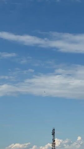 A drone flies through a blue sky filled with scattered clouds. In the foreground, a tall cell tower is visible.