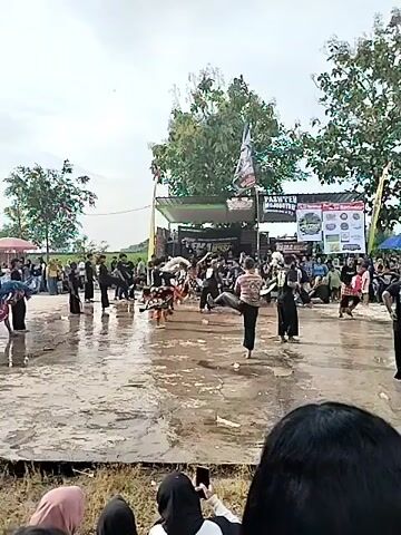 The video shows a performance in what appears to be a wet, open space. Numerous people are present, some dressed in what seem to be traditional or performance costumes, and they are dancing or moving around. Other people, potentially spectators, are observing the event. Banners and signs can be seen in the background, suggesting a specific event or organization. The setting appears to be a rural area, possibly in Southeast Asia, given the style of dance and dress.