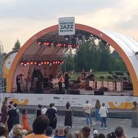 A band is performing on a stage at the Moscow Jazz Festival. The stage is under a large orange arched tent with lights. The band members are playing instruments like a piano, drums, and possibly a saxophone and guitar. A crowd of people are watching the performance from the ground in front of the stage. The weather appears to be overcast, and trees are visible in the background.