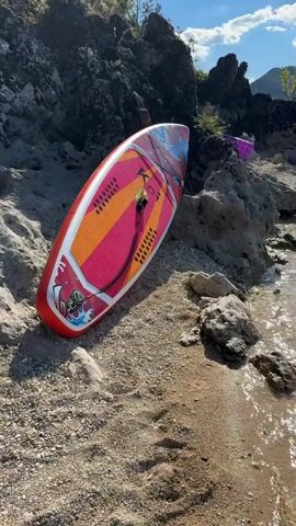 A stand-up paddleboard with a pink, orange, and blue design rests on a sandy and rocky shore. The paddleboard is angled, with the tail facing the water. In the background, a body of water reflects the sunlight, and the shore is lined with rocks and vegetation. Further in the distance, mountains are visible under a blue sky with scattered clouds. A boat is docked on the right side of the frame. The scene appears to be a serene natural environment, possibly a lake or a calm sea.