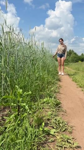A young woman with dark hair, wearing a plaid shirt and denim shorts, walks down a dirt path next to a tall green field of grain under a blue sky with fluffy white clouds. She is smiling and appears to be enjoying the outdoors. The path is well-trodden and bordered by green grass and vegetation. In the distance, more green fields and trees are visible, suggesting a rural or countryside setting. The sun is shining, casting shadows on the path and illuminating the scene.