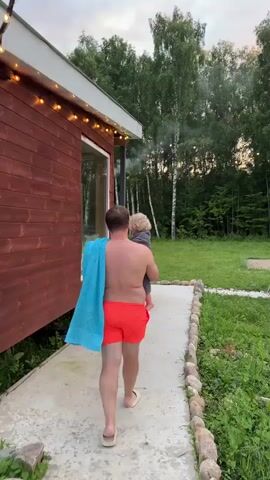 A man wearing red swim trunks and holding a blue towel over his shoulder is walking away from a wooden cabin. He is carrying a small child in his arms. The cabin has a large window and is decorated with string lights. A path made of concrete and lined with stones leads away from the cabin into a grassy area with trees in the background. Smoke is visible rising from a chimney or pipe near the cabin. The sky appears overcast.
