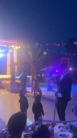 A night-time outdoor party is underway with a stage set up at the back. Multiple people are present, some dressed in costumes, including one individual in an elephant costume and another in a long black suit with a hat and cane. A few children are watching the festivities. Colorful lights illuminate the area, and flags are strung across the top of the scene. The overall atmosphere suggests a festive event, possibly a celebration or a performance.