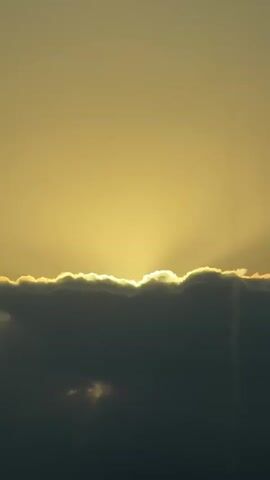 The video shows a time lapse of a sunrise through clouds. The sun peeks over a dark cloud formation, casting rays of light into the sky. The sky is a pale yellow color, and the clouds are dark gray with a white, illuminated edge. As the time lapse continues, the sun rises higher, and the clouds begin to thin, revealing a landscape with hills and trees in the distance. The video ends with a close-up shot of the sun's rays breaking through the clouds.