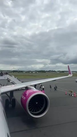 The video captures an exterior view from inside an airplane, showcasing the boarding process on an airport tarmac. The dominant feature is a Wizz Air aircraft, identifiable by its bright pink and white livery. In the foreground, the large pink engine nacelle is prominent, displaying "wizzair.com" in white text. Part of the aircraft's wing stretches across the frame to the right, with the pink tailfin visible in the distance, partially showing the airline's logo.

Numerous passengers, dressed in casual attire and carrying bags, are seen walking across the grey asphalt tarmac. They are moving purposefully towards the front of the aircraft, indicating they are boarding the flight. A path for passengers is marked by orange and white traffic cones connected by ropes.

The weather is overcast, with a heavy grey cloud cover dominating the sky. In the background, beyond the airport apron, there's a flat landscape with distant green areas and some low-rise buildings or airport facilities. A white vehicle is also visible in the distance on the tarmac. The scene takes place during daytime at an airport, specifically in Tillé, France, depicting a routine passenger boarding operation under cloudy conditions.