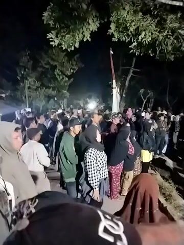 A crowd of people are gathered outdoors at night. They are standing around a small building or structure. Many people are looking in the same direction, suggesting they are watching something. Some individuals are holding up their phones, possibly to record or take pictures. The environment appears to be somewhat rural, with trees and foliage visible. The overall atmosphere suggests a community event or gathering.