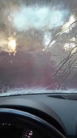 The video shows the view from inside a car, looking out through the windshield. The windshield is wet with water droplets and streaks, suggesting it is raining or being washed. Through the water, a blurry red structure can be seen, possibly part of a car wash. The car's dashboard and steering wheel are partially visible in the foreground. The lighting is dim and diffused, with some bright areas suggesting an external light source obscured by the water and the structure. The overall impression is of being inside a vehicle moving through a wet and possibly enclosed environment.