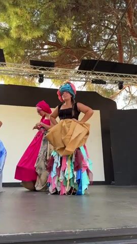 Two women are performing on an outdoor stage. The woman in the foreground is wearing a black top and a skirt made of various colored fabrics and a large piece of burlap fabric draped around her waist. She also has a colorful headdress. She is smiling and has her hands on her hips. The woman in the background is wearing a bright pink dress and a pink headscarf. She has her hand to her face and appears to be looking down. Behind the women, there is a black backdrop and some lighting equipment. Trees are visible above the stage.