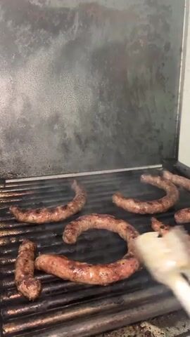 A close-up shot shows sausages being cooked on a grill. The grill, made of metal bars, contains multiple sausages of varying sizes and shapes. A hand holding a pair of metal tongs is visible, manipulating one of the sausages. Smoke rises from the grill, indicating active cooking. The background shows the inside of the grill lid, which has a mottled, metallic appearance. The setting seems to be outdoors, possibly on a patio or deck.