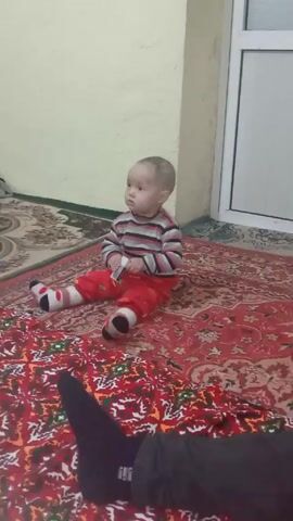 A baby is sitting on a rug in what appears to be an indoor room. The baby is wearing a striped long-sleeved shirt and red pants with red and white socks. The baby is holding a small rectangular object in their hands. An adult walks into the frame, crouches, and then appears to crawl away. The rug is red with intricate patterns. The walls are light yellow. There is a white door or frame in the background.