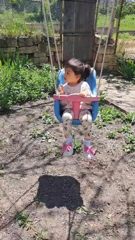 A little girl is sitting in a swing in a backyard on a sunny day. She is wearing a light-colored sweater, leggings with bee designs, white sneakers with pink bows, and white socks. The swing is blue and pink. The backyard has grass, stone walls, a wooden structure, and some plants. The girl is playing with something in her hand and interacting with the camera. She is not being swung.