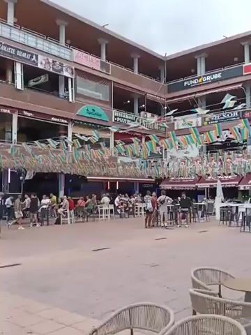 Here's a concise description of the video:

The video shows a bustling outdoor plaza, likely in a tourist area. It's a paved area with multiple restaurants and bars surrounding it, situated in a multi-story building complex. Many people are milling around, some sitting at tables outside the establishments, others walking through the area. The plaza is decorated with colorful banners and flags strung overhead, giving it a festive atmosphere. Signs for various restaurants and bars are visible, with names like "Wapa Tapa," "Namaste Bharat," and "Flamingo." The overall impression is of a vibrant and busy location, possibly a popular spot for dining and socializing.