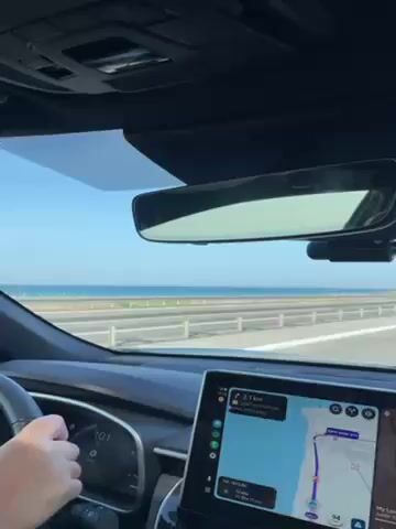 A car is moving along a highway on a bright, sunny day.  The ocean is visible to the left of the highway. The car's navigation system shows a map. The dashboard is visible and a hand is on the steering wheel. A pink air freshener is visible attached to an air vent. The sky is blue with very few clouds. Other vehicles are visible on the highway. A "Toyota Relax" sticker is on the windshield.