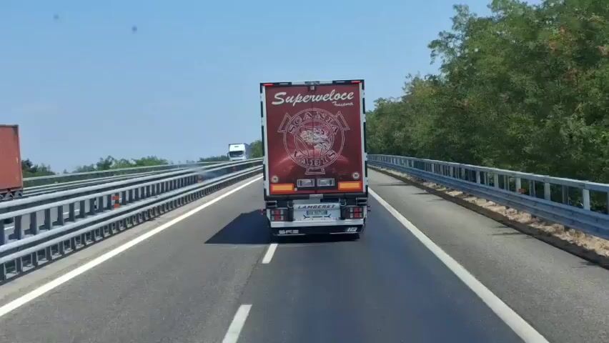 The video shows a view from a vehicle driving on a highway. The main focus is a large red truck with "Superveloce Trasporti" written on the back. The truck has a circular logo with text and an image in the center. There are other trucks in the distance on the highway, and trees and vegetation line the sides of the road. The sky is blue and clear, indicating a sunny day. The highway has guardrails on both sides.