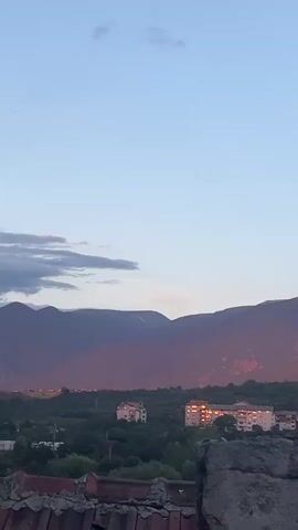 A vertical, low-angle shot looks out over rooftops towards distant mountains. The sky is a pale blue with scattered clouds. The mountains are in shadow, with the setting sun casting a warm, reddish-pink glow on their peaks. Below the mountains, a dense green forest is visible, dotted with some buildings. In the foreground, the tiled roofs of several houses and a concrete chimney are prominent. The image appears to be taken at dusk or dawn.