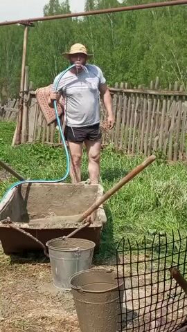 The video shows a man in a rural setting using a hose to spray water into his mouth and then into a wheelbarrow filled with cement. The man, wearing a straw hat, gray t-shirt, and black shorts, stands in a yard with muddy feet. A blue hose is looped around his neck and extends to his mouth. He sprays water from his mouth into the wheelbarrow. The wheelbarrow contains a gray mixture of cement. Two metal buckets sit in front of the wheelbarrow, also filled with a gray liquid, likely water. A wooden fence surrounds the yard. The background features green trees and grass, suggesting a summer day. Two shovels lean against the wheelbarrow.