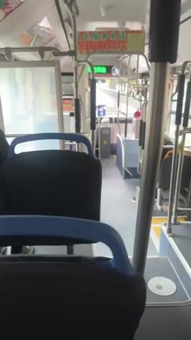 This video appears to be filmed from the interior of a moving bus. The camera pans across the aisle, showing rows of dark seats with blue trim. Several passengers are visible. In the foreground, a person's arm and hand are visible, holding a colorful candy bar. Further down the aisle, a man with a bald head is seated, looking out the window. Another passenger, a younger man wearing a baseball cap, is looking at his phone. Outside the windows, scenery and other vehicles can be seen, indicating the bus is in motion on a road. There are overhead handrails and advertisements displayed inside the bus.