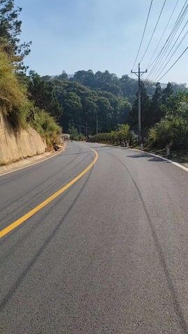 The video shows a grey asphalt road winding through a rural environment. A solid yellow line marks the center of the left lane. There are tire tracks visible on the road surface. A concrete barrier runs along the left side of the road, while the right side has a grassy shoulder. Utility poles line the right side of the road. The road curves uphill and out of sight. Lush green trees cover the surrounding hills. The sky is clear and blue. A small figure on a motorcycle can be seen in the distance along the road.