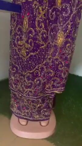The video shows a headless mannequin wearing a purple dress with gold embroidery and red accents. The dress is made up of a fitted bodice with puffy short sleeves and a wrap skirt with a ruffled detail at the hip. The mannequin is standing on a light pink base, which in turn is on a green mat. The background includes a white wall, with a shelf holding spools of thread. There is a piece of white paper with red writing on it, sitting on the green mat next to the mannequin. The camera moves up and down the mannequin, showing the full dress. There is music playing in the background.