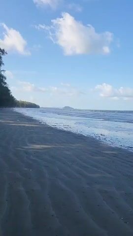 A serene beach scene unfolds under a partly cloudy blue sky. The foreground is dominated by the dark, rippled sand of the beach, showing the marks of receding waves. The ocean stretches out in the middle ground, its surface calm with gentle waves lapping at the shore. In the distance, a lush green island with rolling hills rises from the sea. The overall impression is one of peaceful natural beauty.