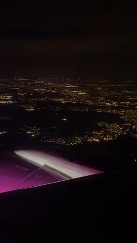The video shows a night cityscape from an airplane window. The city is illuminated by numerous lights, appearing as a vast network of yellow, orange, and red dots spread across a dark landscape. The wing of the plane is visible in the foreground, with a white and purple light shining on the section closest to the viewer. The text "NO STEP" is visible on the wing. The sky above is dark and cloudy, with a few faint stars visible. The camera is relatively stable, offering a clear view of the city below.