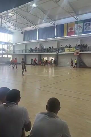 The video shows an indoor futsal game. The court is made of wood with white lines marking boundaries and areas. A futsal ball is visible on the court. Several players, dressed in athletic attire, are participating in the game. Spectators are seated in elevated bleachers overlooking the court. The environment appears to be a sports hall with large windows providing natural light and a high ceiling with visible beams and lighting fixtures. Flags are hanging from the upper level of the hall. The overall scene depicts a sporting event in progress.