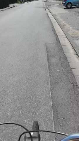 The camera is mounted on a bicycle, showing a first-person view of someone cycling down a paved road. The road has a curb and sidewalk on the right. There's a light-colored car parked on the right side of the road in the distance. The road appears to intersect with another street ahead, indicated by white lines painted on the asphalt. Trees and houses are visible in the background on both sides of the street. The weather is sunny.