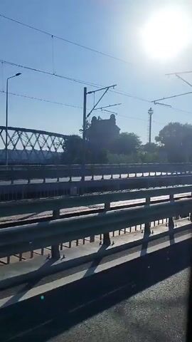 The video shows a view from a moving train. The perspective is from a window, looking out at a landscape. The train is traveling alongside railroad tracks. In the distance, there is a large metal bridge structure that appears to be a railway bridge. To the right of the bridge, there are trees and foliage. Further along, there are some monuments or structures, one of which resembles a tall tower. The sky is clear and blue, with overhead power lines visible, indicating the train is likely electric. The sun is shining, casting shadows from the guardrail in the foreground. The motion of the train creates a sense of movement and passing scenery.