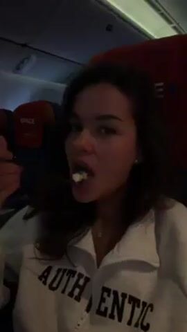 The video shows a woman seated on an airplane. She has light brown hair and is wearing a white sweatshirt with the word "AUTHENTIC" on it. She eats something from a spoon, then she holds a disposable black stirring stick. The airplane interior is visible, with red seat covers labeled "SPACE." The lighting in the cabin appears dim.