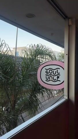 The video shows a street view through a window. Outside, there is a palm tree blowing in the wind, buildings and a light-colored car driving by. A pink sign with the words "SOUL VAUlT" in black is affixed to the window frame. The sky is a pale blue.