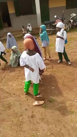 Children are playing a game outdoors. The ground is dirt and some scattered rocks. A building is in the background. A motorcycle is parked near the building. One child is wearing a red hijab and a dark colored dress, and green pants. Another child is wearing a blue and white outfit, and khaki pants. A child in the foreground is wearing a white shirt and bright green pants, with an orange headscarf. There are two brown sandals on the ground in front of this child. Another child is wearing a white outfit with a white headscarf and green pants. A child is kneeling. The children appear to be engaged in some form of hopscotch or similar game, with a stick in the dirt and possibly drawn squares.