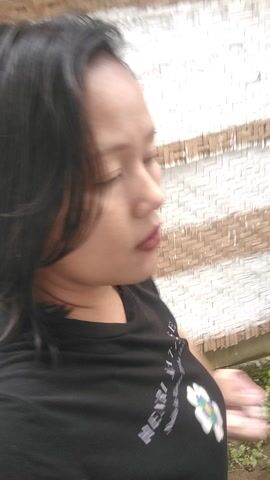 A young woman with dark hair and a neutral expression is featured in a close-up, slightly tilted video. She is wearing a black t-shirt with the words "HENRI MATISSE" and "Nadia Au Viv..." visible on the front. Her lips are adorned with dark red lipstick. The lighting is bright, suggesting it is daytime. The background appears to be an outdoor setting with light-colored, possibly wooden, structures and a bright, somewhat overexposed sky or wall. The woman looks directly into the camera, blinking occasionally and moving her head slightly. The overall impression is a casual, outdoor selfie.