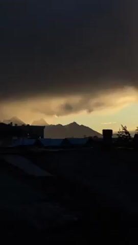 The video shows a view from a roof overlooking other buildings and mountains in the distance. The scene is dark, with a large, dark cloud formation dominating the sky. A golden hue is visible where the clouds are thinner, suggesting the sun is setting or rising. The outlines of the mountains are visible against the lighter sky. The roofs in the foreground are silhouetted and appear to be made of corrugated metal. A few trees are visible on the right side of the frame.