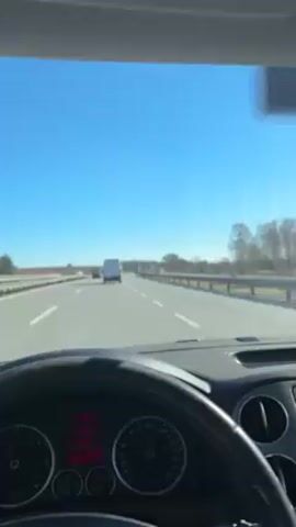 A forward-facing interior shot from the driver's seat of a car moving on a highway during the day. The sky is clear and blue. The road has multiple lanes, and a few other vehicles are visible in the distance. The dashboard, steering wheel, and air vents of the car fill the lower portion of the frame. Roadside barriers are visible to the right of the road. Road signs are seen.